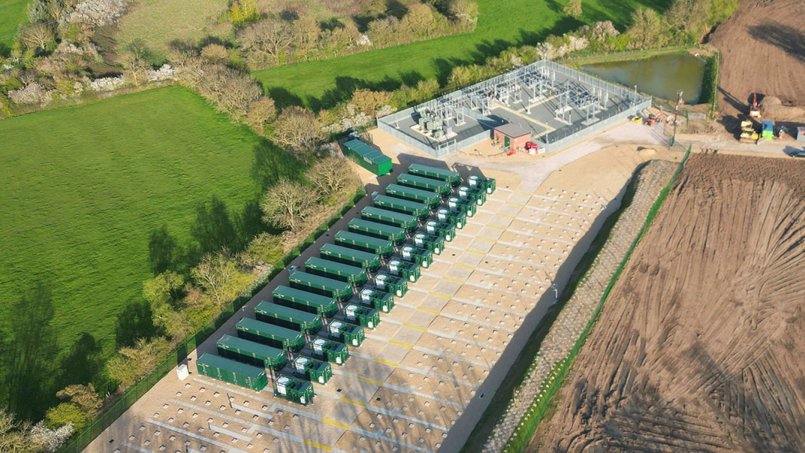 Aerial view of a battery energy storage system (BESS) installation with green containerized battery units and a substation facility, surrounded by green fields and construction areas