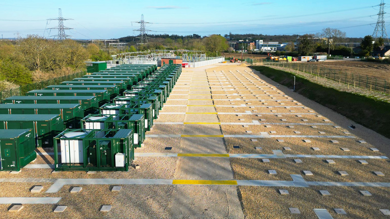Ground-level view of battery energy storage facility featuring multiple green container units and electrical infrastructure on a gravel site, with pylons and industrial buildings visible in the background.