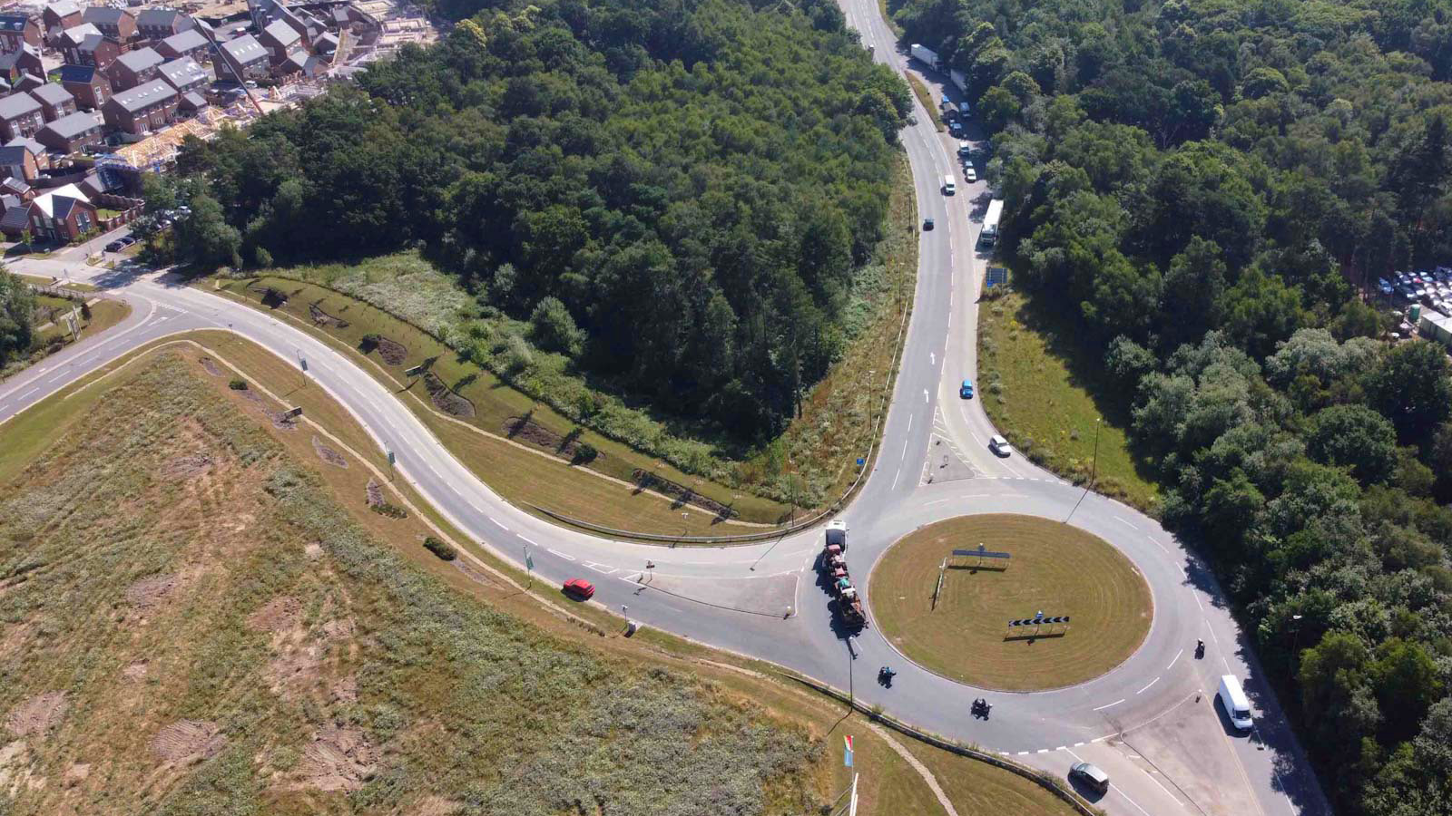Aerial view of a landscaped roundabout connecting to a residential housing development and forested area, with vehicles on a multi-exit road network.