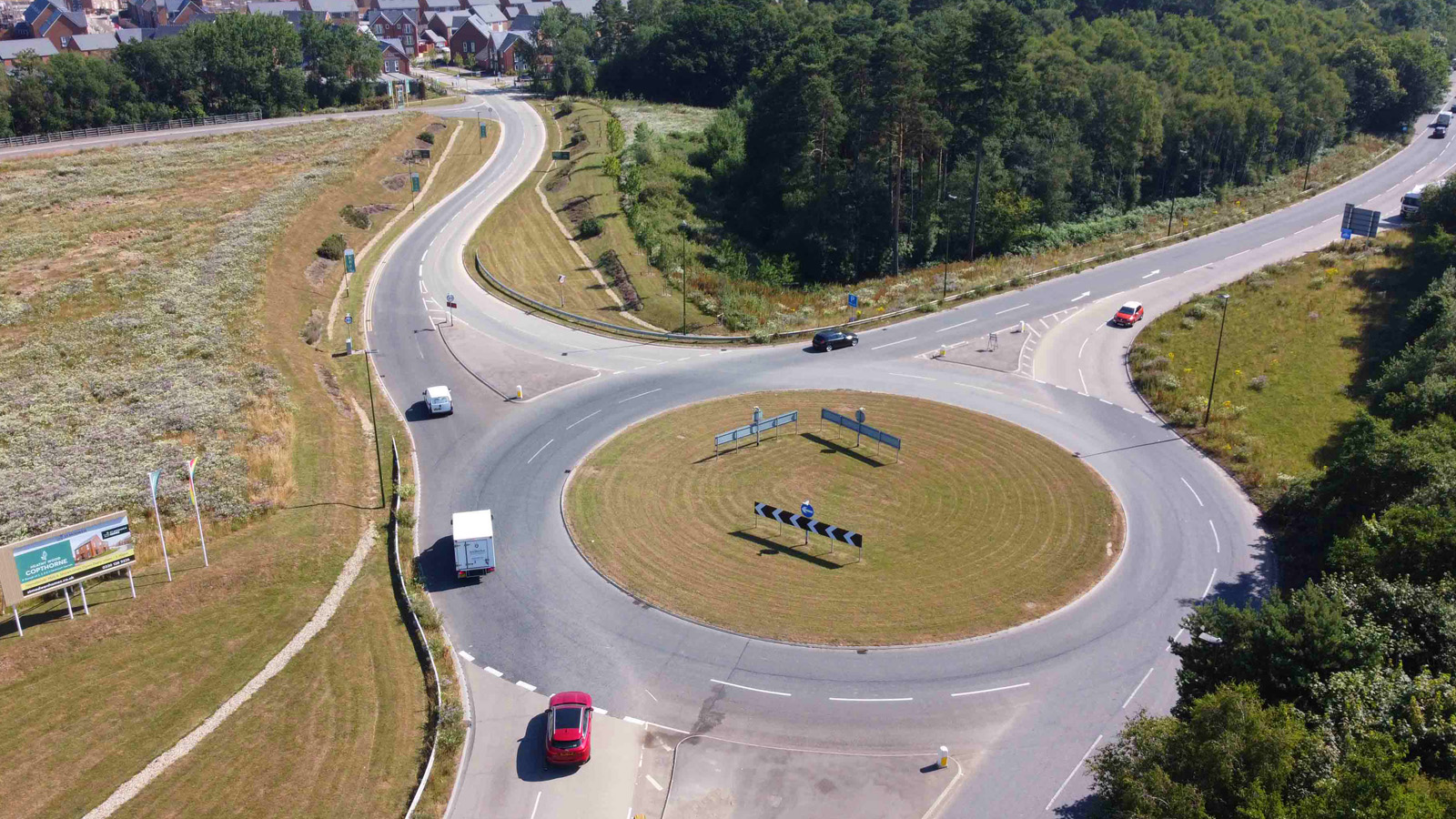 Aerial view of a modern roundabout connecting to a residential housing development, with landscaped verges, open fields, and surrounding woodland