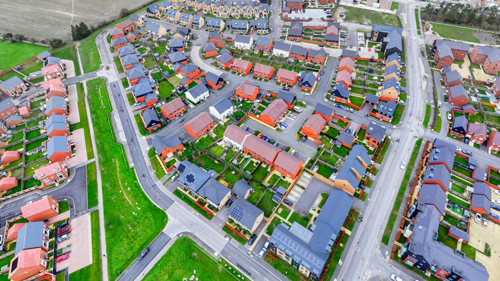 Aerial view of a modern housing development featuring red-brick homes, landscaped gardens, paved roads, and green communal spaces in a suburban area.