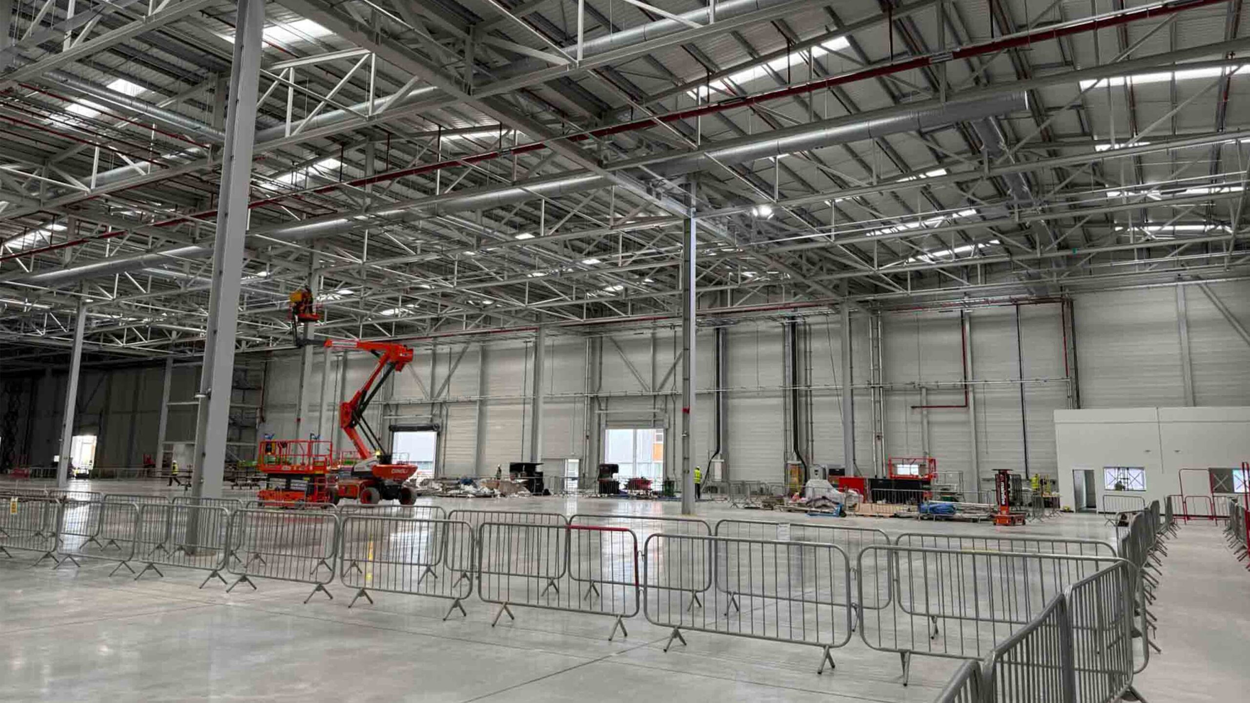 Industrial Warehouse Construction with Steel Framework and High-Level Access | Natta Large industrial warehouse interior under construction with steel framework, high ceilings, and workers using cherry pickers for installation - BMW Mini Plant Oxford - Groundworks Package