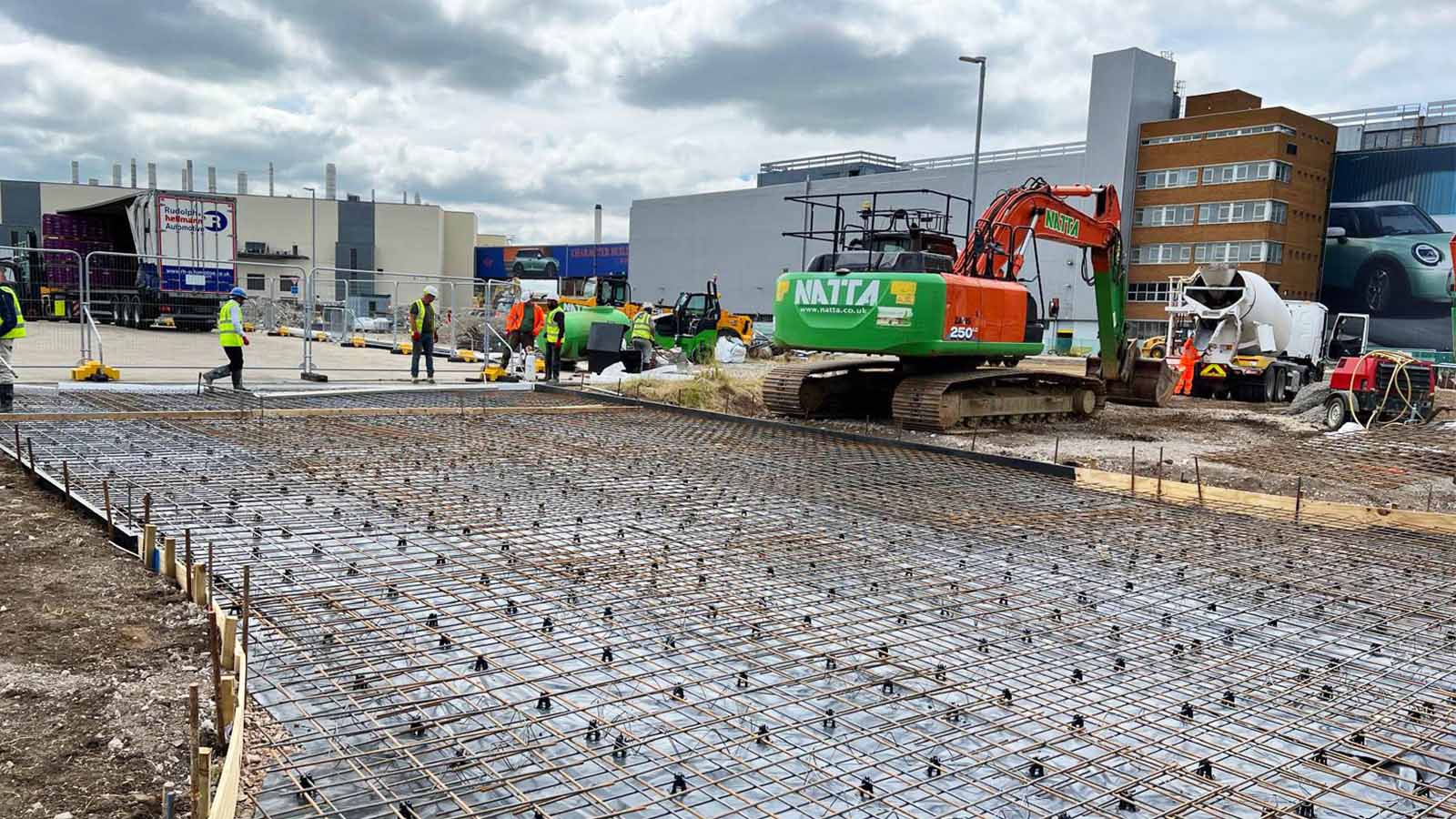 Concrete slab preparation at a construction site in Oxford. | Natta Construction workers preparing reinforced steel mesh for concrete slab pouring, with excavators, a cement mixer, and industrial buildings in the background - BMW Mini Plant Oxford - Groundworks Package
