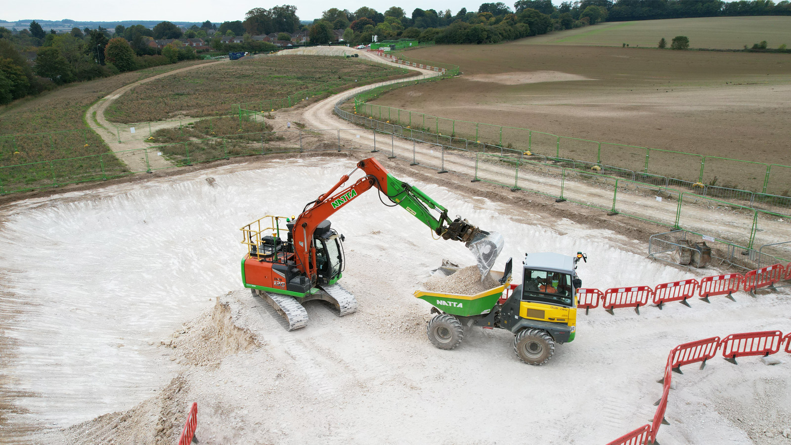 Excavator loading gravel into a dump truck during site excavation at a construction project, surrounded by red safety barriers and rural landscape - S278 Works