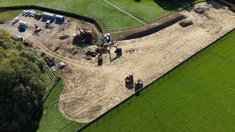 Aerial view of a construction site with dirt roads, heavy machinery, and vehicles inside a fenced area surrounded by green fields and trees.