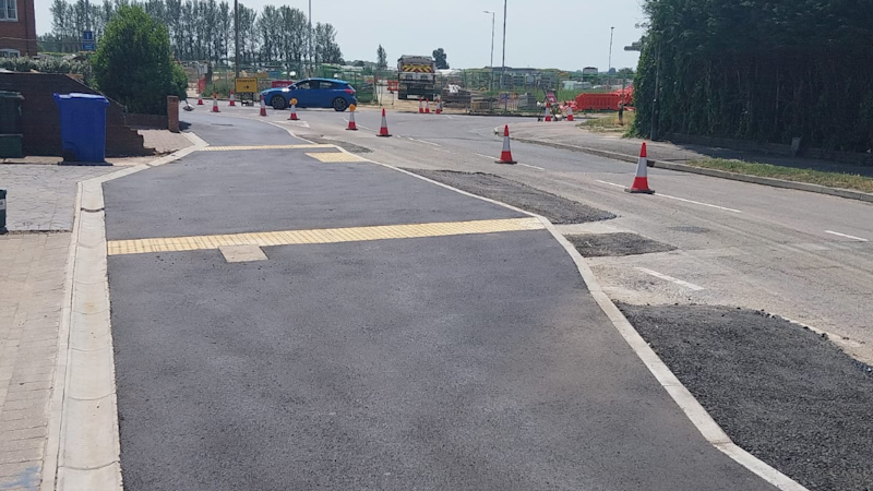 Road under construction with fresh asphalt and traffic cones guiding vehicles; curb and sidewalk on the left, and a blue car in the distance.