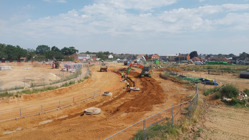 Construction site with a dirt road, excavator and dump truck working, workers guiding soil, and temporary fencing; piles of materials and houses in the distance.