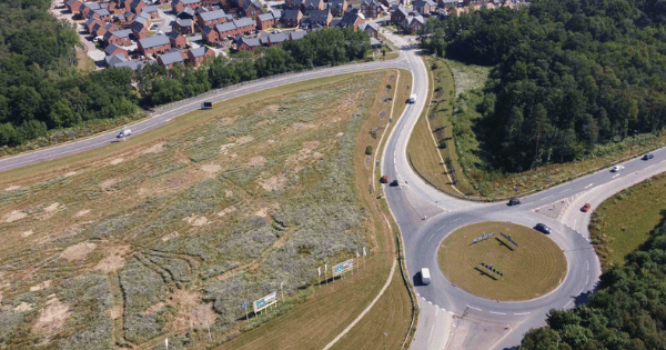 Aerial view of a roundabout connected to a curved road, with a residential area to the top-left and wooded areas on the right.