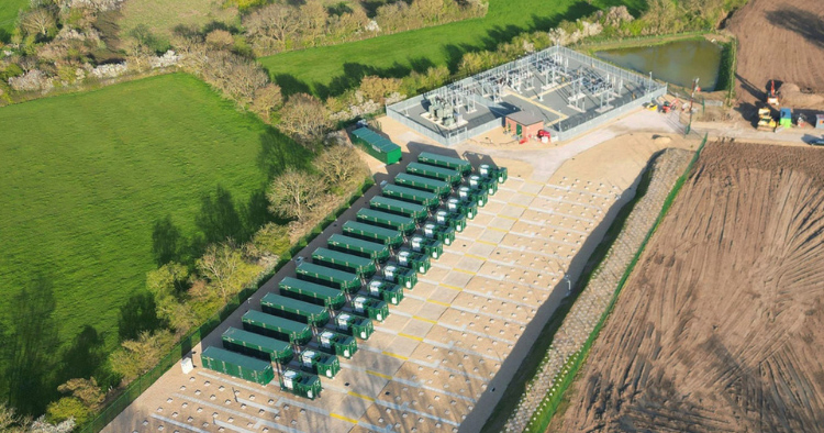Aerial view showing a row of green storage containers or trailers lined up on a paved yard next to a utility substation under construction, with surrounding fields.