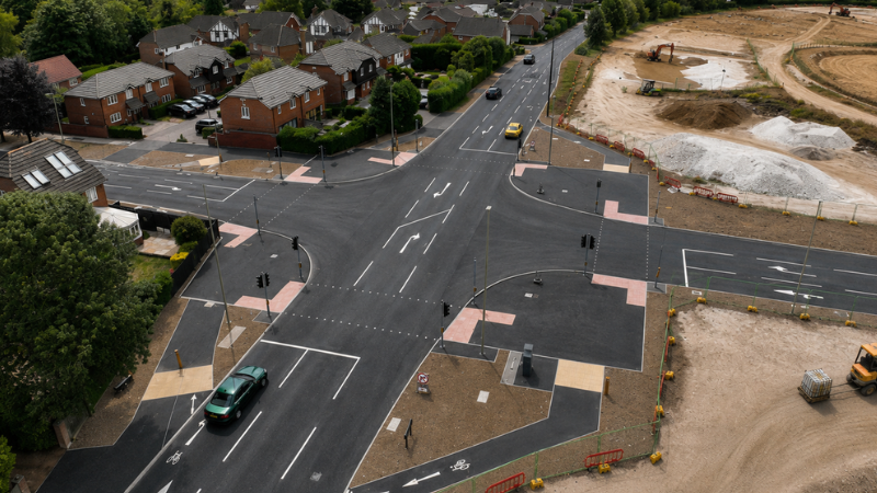 Aerial view of a newly paved urban intersection with traffic lights, crosswalks, and surrounding houses and a construction site nearby.