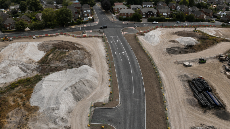 Aerial view of a newly paved curved road under construction flanked by dirt piles and construction equipment, with a residential street and houses in the background.