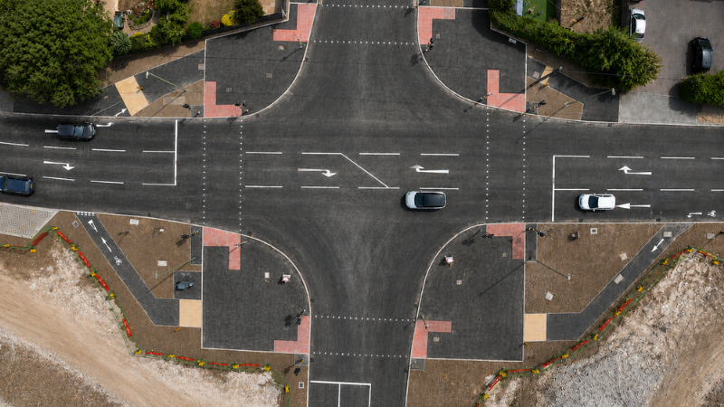 Aerial view of a four-way intersection with curved ramps, lane markings, and several cars traveling on dark asphalt.